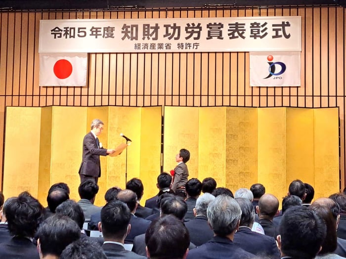 【写真】知財功労賞表彰式の様子（大勢の人の前で表彰状を受け取る代表の垣内）