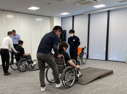 【写真】車いす実技の様子。車いすに乗る受講者をもう一人の受講者が後ろから押しながら、簡易スロープを上る様子。