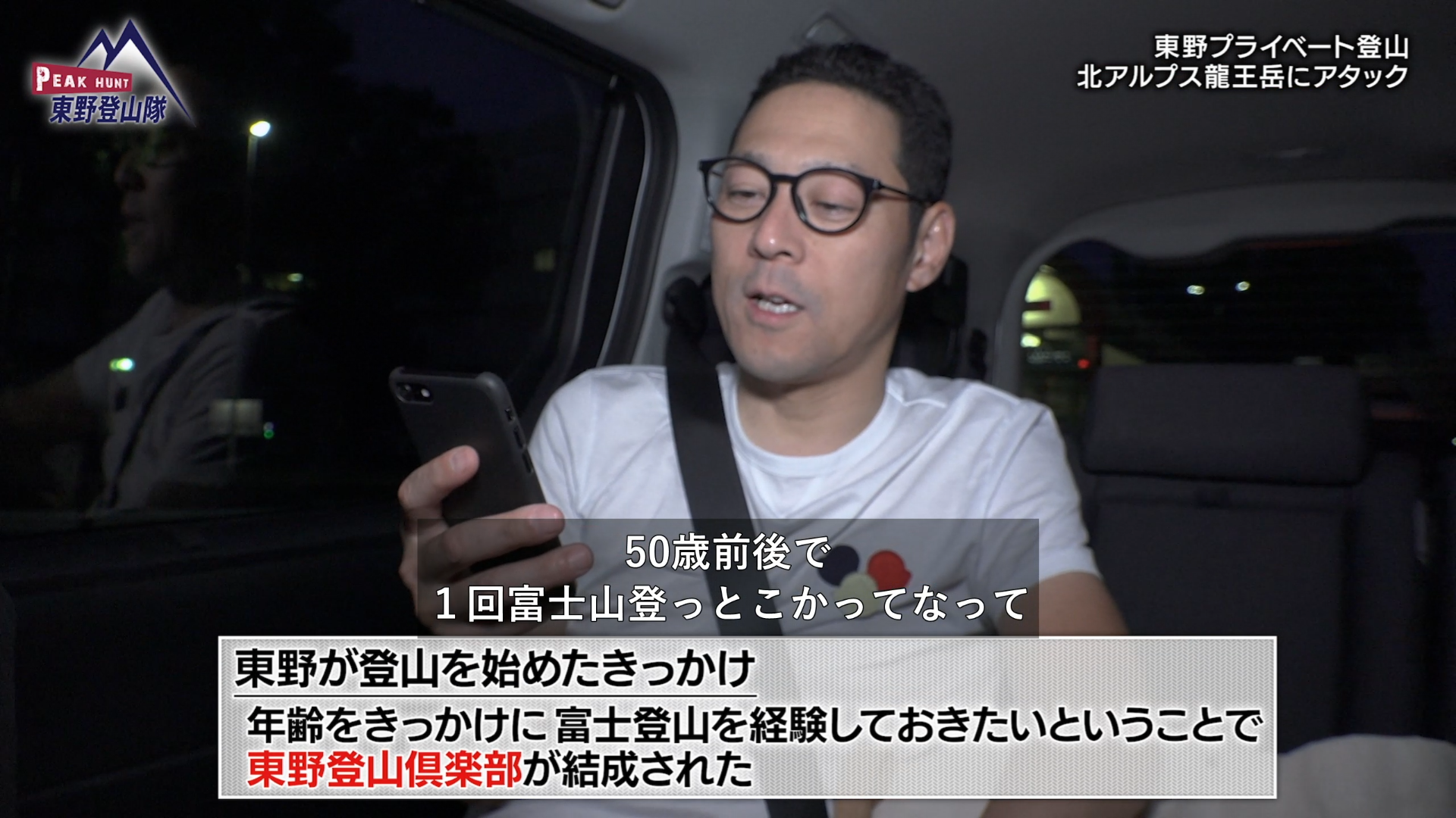 【画像】「東野登山隊」の一場面。東野さんが車に乗ってスマホを触っている様子と「東野が登山を始めたきっかけ、年齢をきっかけに富士登山を経験しておきたいということで東野登山俱楽部が結成された」というテロップ。その上部に「50歳前後で1回富士山登っとこっかなって」という字幕。